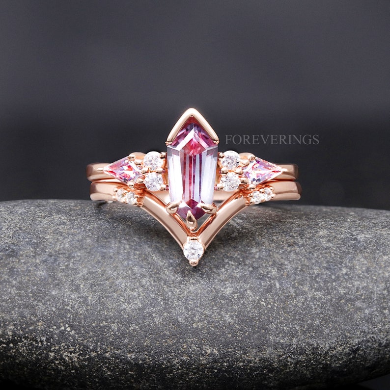 May include: A rose gold engagement ring set with a large, rectangular, purple and pink gemstone surrounded by smaller, round, white gemstones. The ring is set on a gray, textured surface. The text "FOREVERINGS" is visible in the upper right corner of the image.