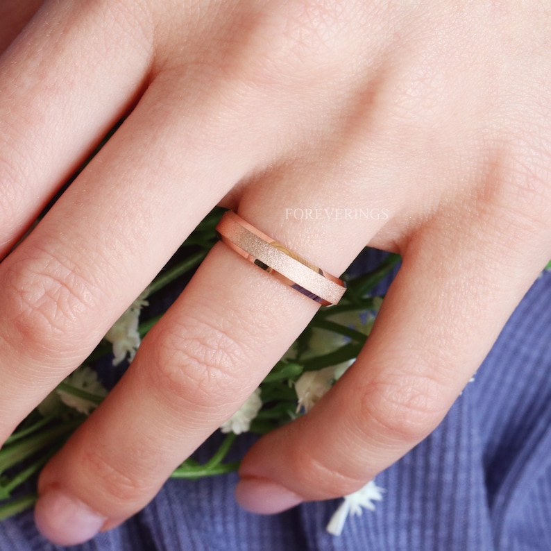May include: A rose gold wedding band with a brushed finish and a beveled edge. The band is worn on a finger.