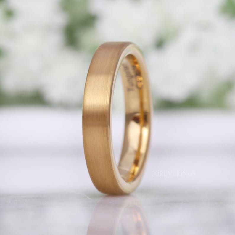 May include: A gold-colored ring with a brushed finish. The ring has a flat profile and a smooth, matte surface. The inside of the ring is a shiny gold color. The ring is likely made of tungsten. The word "Foreverings" is printed on the ring.