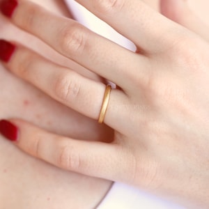 May include: A simple gold band ring worn on a finger. The ring is smooth and has a matte finish.