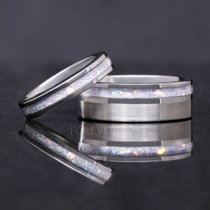 May include: Two silver wedding bands with a thin iridescent glitter band on each. The bands are on a black reflective surface.