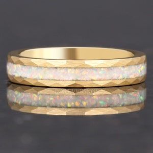 May include: A gold ring with a textured band and a central inlay of white opal. The opal has flecks of color, and the ring is reflected in the surface below.