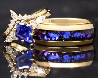 May include: Two gold rings. One ring features a large, blue, emerald-cut gemstone surrounded by smaller white gemstones. The other ring has a gold band with a black inlay filled with blue and purple flecks. The rings are displayed on a reflective surface.
