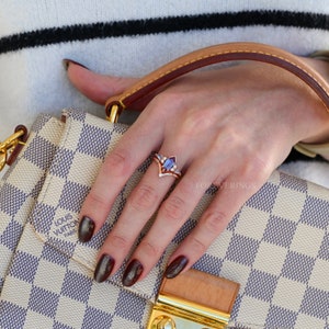 May include: A close-up of a hand wearing a gold ring with a purple gemstone and smaller diamonds. The ring is set in a V-shape with a pointed center. The hand is holding a brown leather handbag with a white and brown checkered pattern. The handbag has the Louis Vuitton logo and the text "Paris" on it.