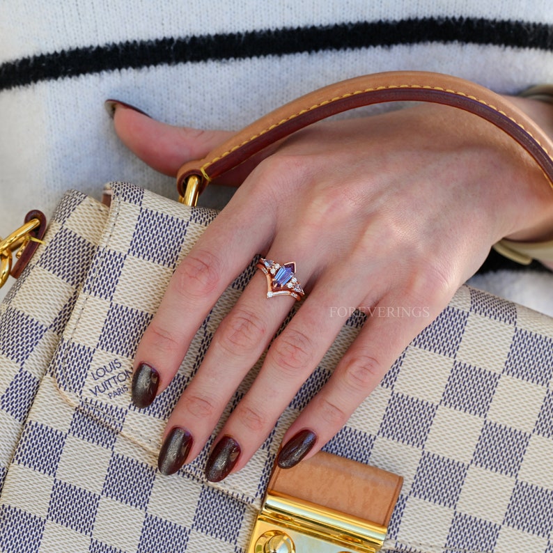 May include: A close-up of a hand wearing a gold ring with a purple gemstone and smaller diamonds. The ring is set in a V-shape with a pointed center. The hand is holding a brown leather handbag with a white and brown checkered pattern. The handbag has the Louis Vuitton logo and the text "Paris" on it.