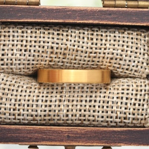 May include: A gold-colored ring with a flat, brushed surface rests inside a wooden box lined with tan burlap fabric. The ring is centered in the frame, showcasing its simple, elegant design. The box is open, revealing the ring's placement.