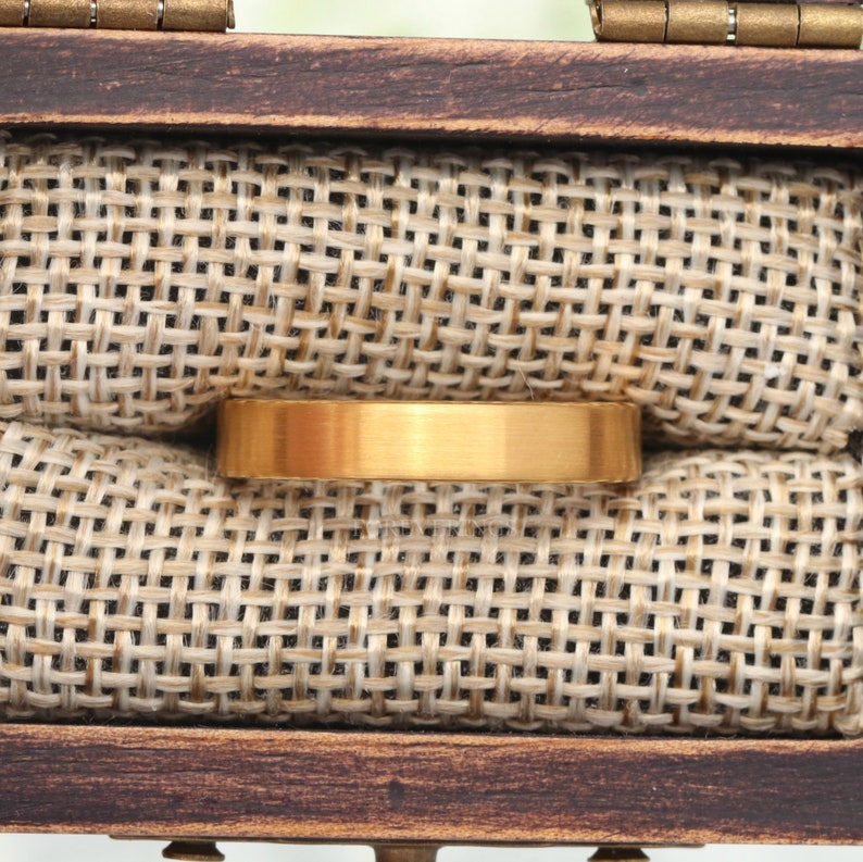 May include: A gold-colored ring with a flat, brushed surface rests inside a wooden box lined with tan burlap fabric. The ring is centered in the frame, showcasing its simple, elegant design. The box is open, revealing the ring's placement.