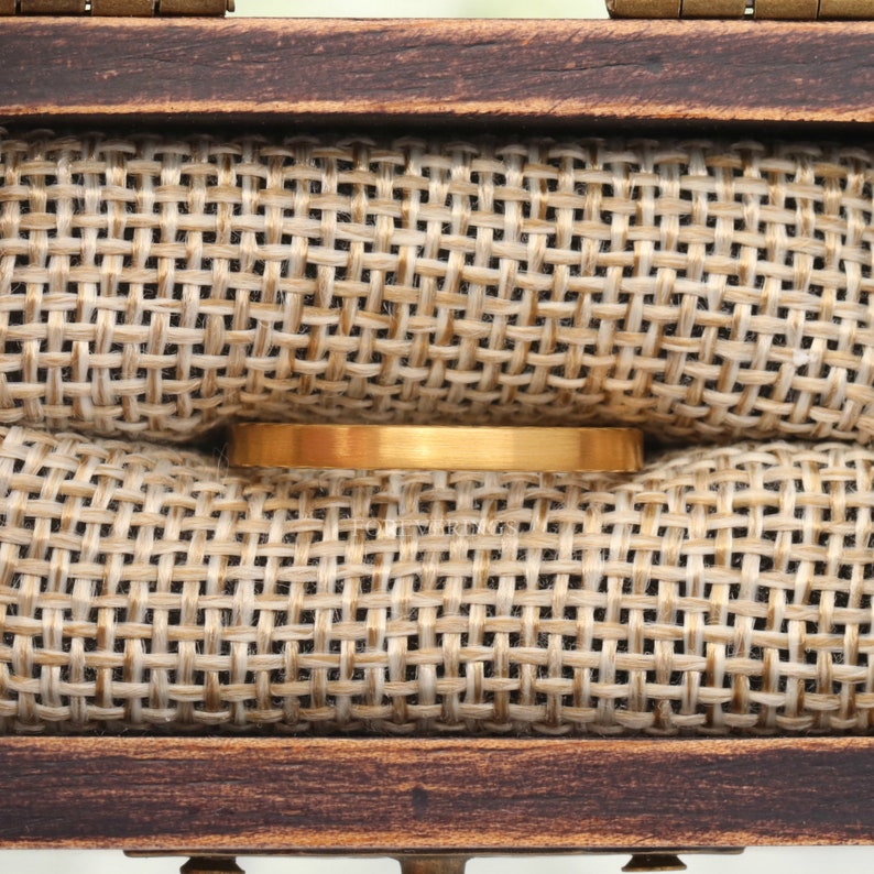 May include: A gold-colored ring displayed inside a wooden box with a burlap-textured interior. The ring is rectangular and has a matte finish. The box has a dark brown, rustic appearance.