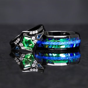 May include: A black wedding band set with a green gemstone and blue accents. The band is made of black tungsten carbide and features a blue and green opal inlay. The inscription "FOREVERINGS" is visible on the band.