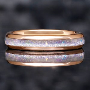 Könnte beinhalten: Roségoldener Ring mit einem Band aus schimmerndem, weißem Opal. Der Opal hat schillernde Farbflecken. Der Ring wird auf einer reflektierenden Oberfläche gezeigt.