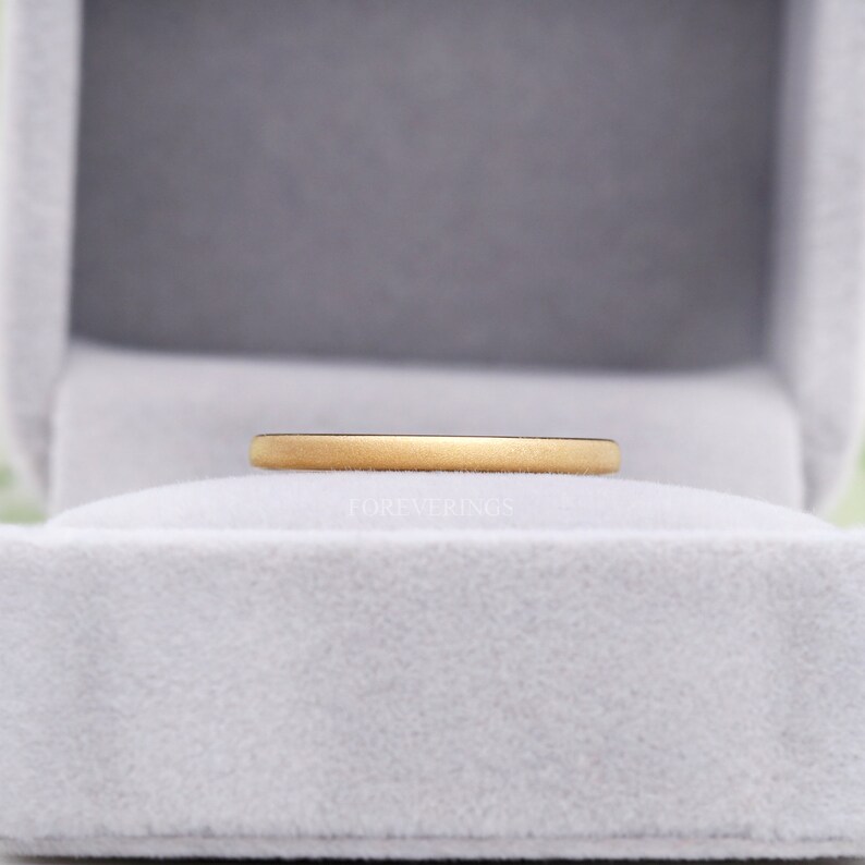 May include: A simple gold wedding band in a white velvet ring box. The band is smooth and has a matte finish.