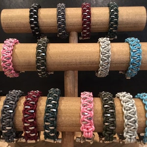 May include: A display of 14 colourful braided bracelets with silver metal accents. The bracelets are displayed on a wooden stand.