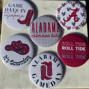 May include: A collection of white, red, and gray buttons with various designs and text. The buttons feature phrases like "Game Days in Tuscaloosa," "Alabama Crimson Tide," and "Roll Tide." Other buttons display a cowboy hat, the letter "A," and cowboy boots.