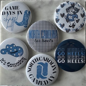 Könnte beinhalten: Eine Sammlung runder Buttons mit verschiedenen Designs und Texten. Die Buttons zeigen Sätze wie "Game Days in Chapel Hill", "North Carolina Tar Heels" und "Go Heels!" in Blau und Weiß. Einige Buttons haben Cowboy-Stiefel- und Hut-Grafiken.