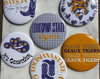 May include: A selection of six circular badges with different designs and text. The badges display phrases such as "Game Days in Baton Rouge" and "It's Gameday", alongside images of a cowboy hat, boots, and a tiger. The color palette includes white, blue, and gold.