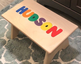 Puede incluir: Un taburete de madera con el nombre "HUDSON" escrito en piezas de rompecabezas de colores. Las letras son de colores arcoíris, cada letra de un color diferente.