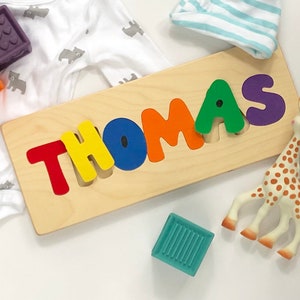 May include: A wooden puzzle with the name "THOMAS" spelled out in colorful letters. The letters are red, yellow, blue, orange, green, and purple. There are also some colorful baby toys in the image.