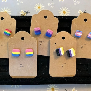 Könnte beinhalten: Sechs Paare quadratischer Ohrringe auf Karten. Die Ohrringe sind aus Polymer Clay gefertigt und zeigen verschiedene Pride-Flaggen-Designs, darunter Regenbogen, bisexuell, transgender und pansexuell.