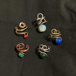 May include: Six ear cuffs with different coloured stones. The ear cuffs are made of wire and have a spiral design. The stones are red, blue, green, and white.