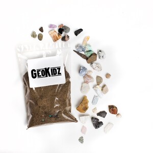 Pode incluir: Um saco de pl&aacute;stico transparente cheio de terra marrom e um r&oacute;tulo branco que diz "GEOKIDZ". Espalhadas ao redor do saco est&atilde;o v&aacute;rias pedras coloridas e marrons.