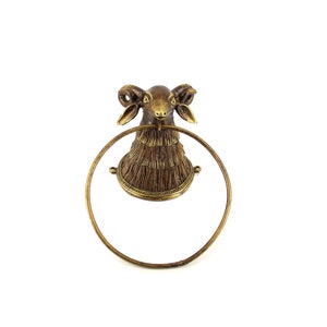 Puede incluir: Un toallero de color bronce con un diseño de cabeza de carnero. La cabeza de carnero tiene cuernos detallados y un aspecto texturizado. El anillo es circular y está hecho del mismo metal de color bronce. El toallero está montado sobre un fondo blanco.