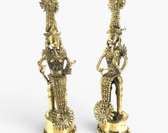 May include: Two tall, metallic gold-toned brass figurines standing on cylindrical bases. Each figure features an elaborate, tall headdress, detailed textured clothing, and circular decorative elements integrated into the base and attire.
