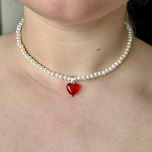 May include: A pearl choker necklace featuring a small, red heart pendant. The necklace is made of small, round pearls. The heart pendant is a bright red, creating a striking contrast against the white pearls. A classic and elegant piece.
