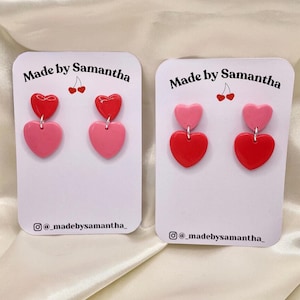 May include: Two pairs of handmade polymer clay earrings. Each earring features two heart shapes, one pink and one red. The earrings are displayed on white cards with the text "Made by Samantha" and the social media handle "@madebysamantha_" printed on them.