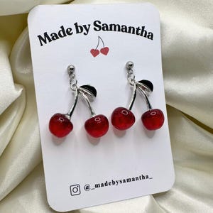 Puede incluir: Pendientes de cereza con cerezas de resina roja, tallos plateados y hojas negras. Los pendientes se muestran en una tarjeta blanca con el texto "Made by Samantha" y un identificador de redes sociales.