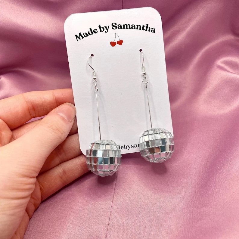 May include: A pair of silver disco ball earrings with a long, thin silver chain. The earrings are displayed on a white card with the text "Made by Samantha" and a small red cherry graphic.