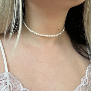 May include: A pearl choker necklace. The necklace is made up of small, round, white pearls, strung together to create a close-fitting collar. The pearls have a subtle sheen, and the necklace sits elegantly against the skin.