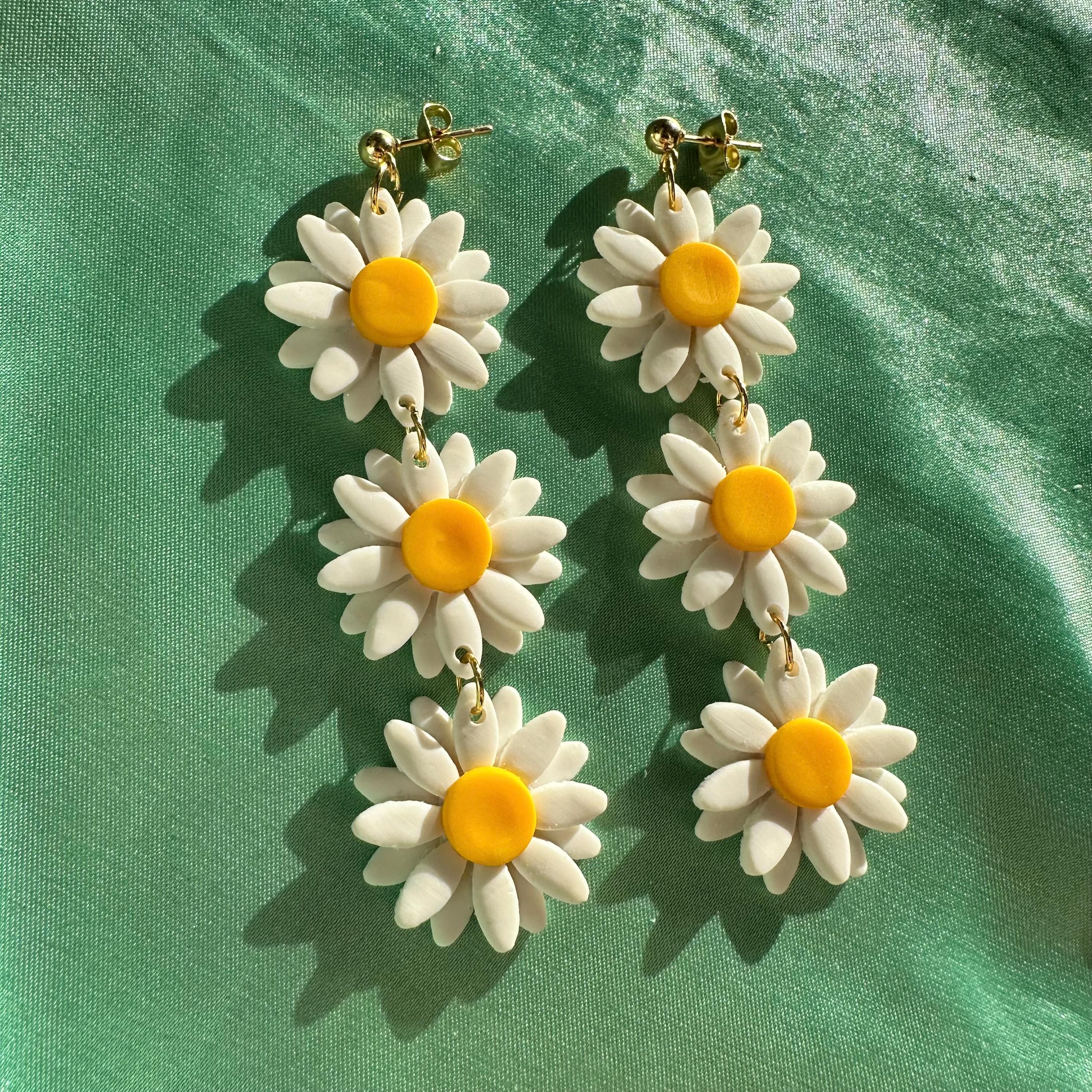 May include: A pair of long white daisy earrings with yellow centers. The earrings are made of polymer clay and have a gold hook.