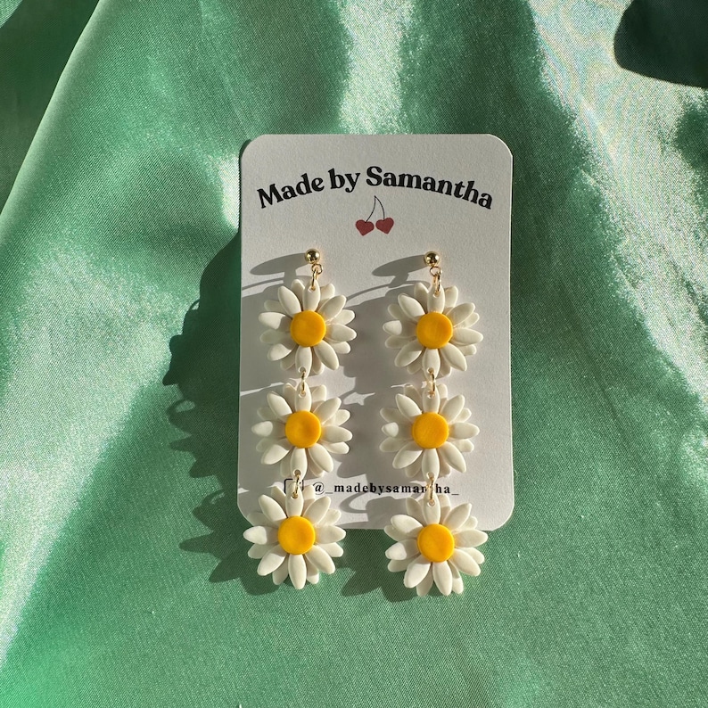 May include: A pair of white daisy earrings with yellow centers. The earrings are made of polymer clay and are strung on gold hoops. The earrings are displayed on a white card with the text "Made by Samantha" and a cherry graphic.