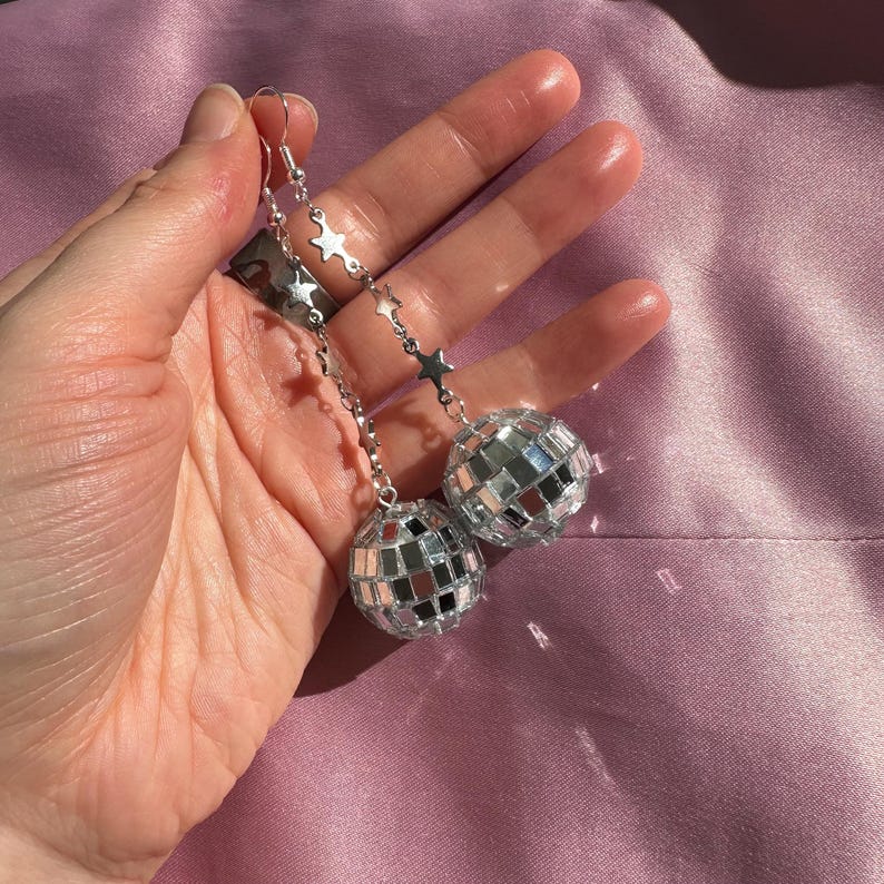 May include: Silver disco ball earrings with a star chain. The earrings are dangling from the earlobes.