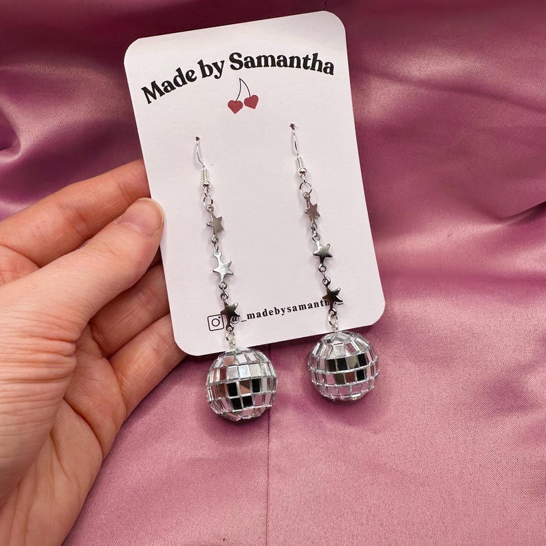 May include: Silver disco ball earrings with a star chain. The earrings are on a white card with the text "Made by Samantha" and a cherry graphic.