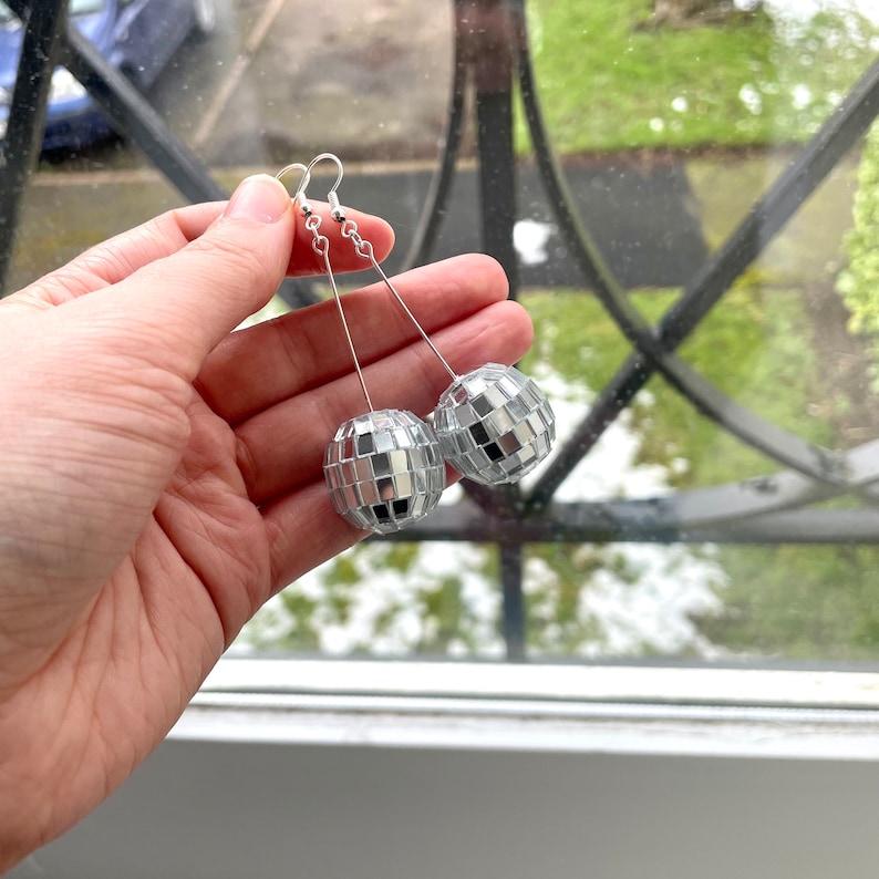 May include: A pair of silver disco ball earrings with a dangling design. The earrings are held in a hand against a window background.