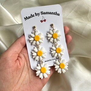 May include: White daisy earrings with yellow centers. The earrings are made of polymer clay and have a gold hook.