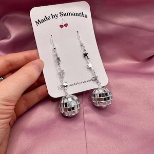 May include: Silver disco ball earrings with a chain of silver stars. The earrings are on a white card with the text "Made by Samantha" and a small red cherry graphic.