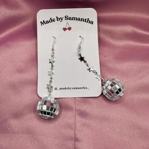 May include: Silver disco ball earrings with a star chain. The earrings are on a white card with the text "Made by Samantha" and "@madebysamantha_" printed on it.
