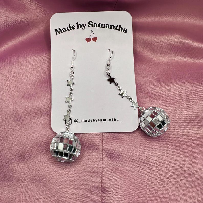 May include: Silver disco ball earrings with a star chain. The earrings are on a white card with the text "Made by Samantha" and "@madebysamantha_" printed on it.