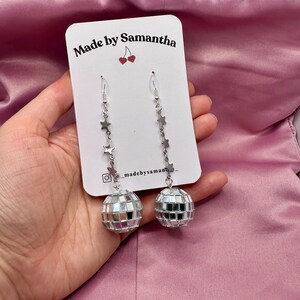 May include: Silver disco ball earrings with a star charm chain. The earrings are on a white card with the text "Made by Samantha" and a cherry graphic.
