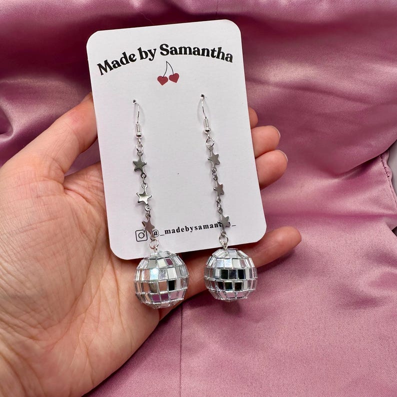 May include: Silver disco ball earrings with a star charm chain. The earrings are on a white card with the text "Made by Samantha" and a cherry graphic.