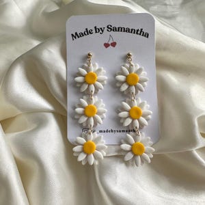 May include: A pair of white and yellow daisy dangle earrings with three daisies on each earring. The earrings are on a white card with the text "Made by Samantha" and a small red cherry graphic.