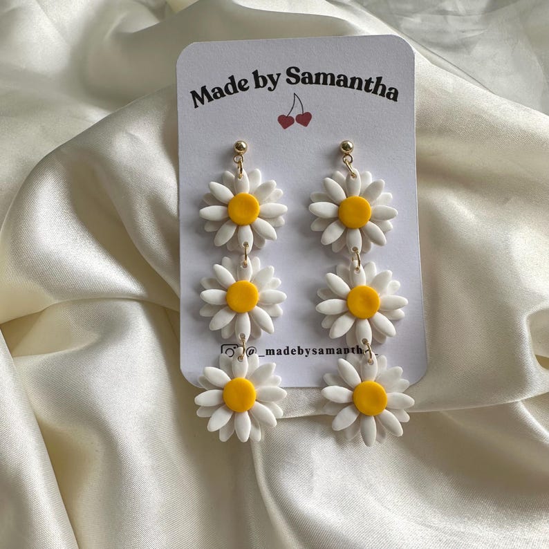 May include: A pair of white and yellow daisy dangle earrings with three daisies on each earring. The earrings are on a white card with the text "Made by Samantha" and a small red cherry graphic.