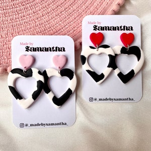 May include: Two pairs of handmade earrings featuring a cow print heart design. One pair has pink hearts and the other has red hearts. The earrings are on white cards with the text "Made by Samantha" and an Instagram handle.