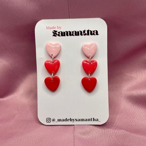 May include: A pair of dangle earrings with three hearts each. The top heart is pink, the middle heart is red, and the bottom heart is red. The earrings are on a white card with the text "Made by Samantha" and "@madebysamantha".