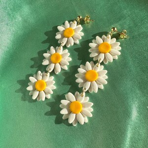 May include: A pair of dangle earrings featuring three white daisy flowers with yellow centers on each earring. The earrings are connected by gold chains.