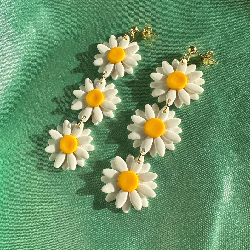 May include: A pair of dangle earrings featuring three white daisy flowers with yellow centers on each earring. The earrings are connected by gold chains.