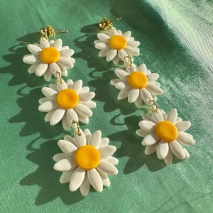 May include: A pair of dangle earrings featuring three white daisy flowers with yellow centers. The earrings are connected by gold jump rings.