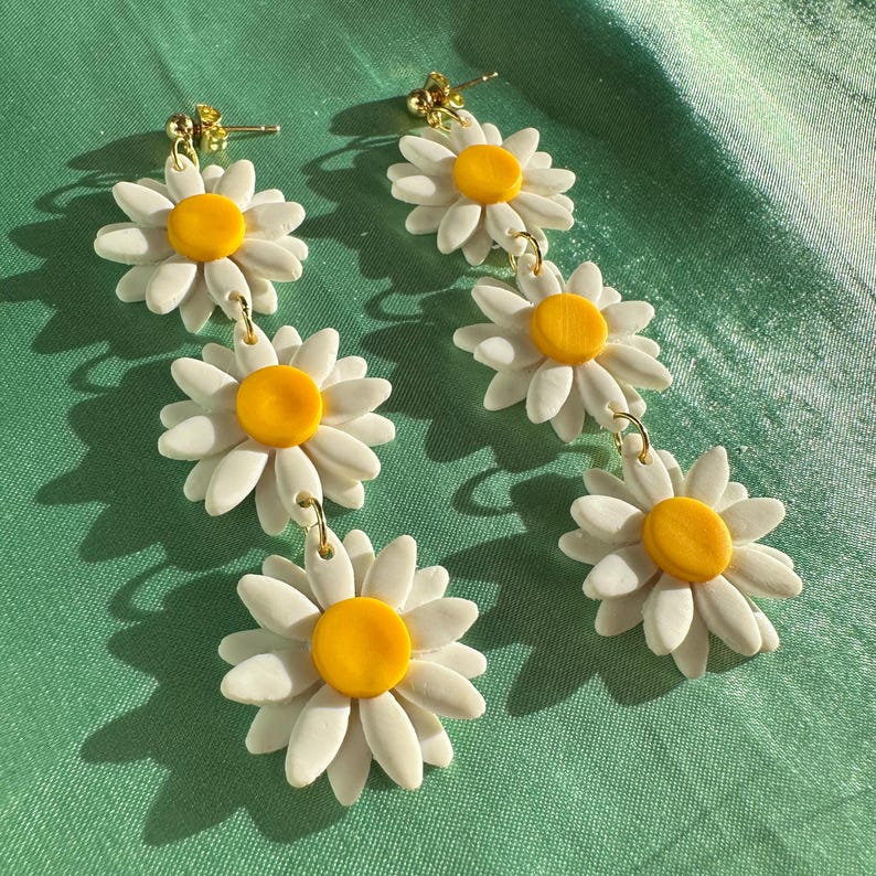 May include: A pair of dangle earrings featuring three white daisy flowers with yellow centers. The earrings are connected by gold jump rings.